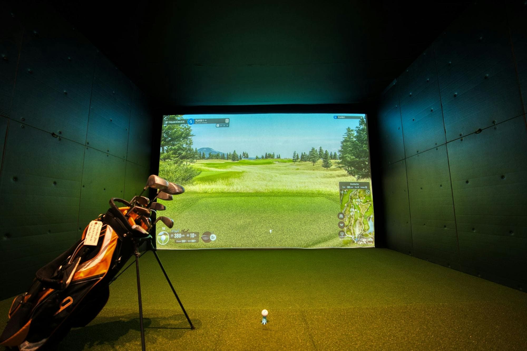 A residential indoor golf simulator bay — a projected course on a hitting screen, club bag on the turf, warm room finishes.